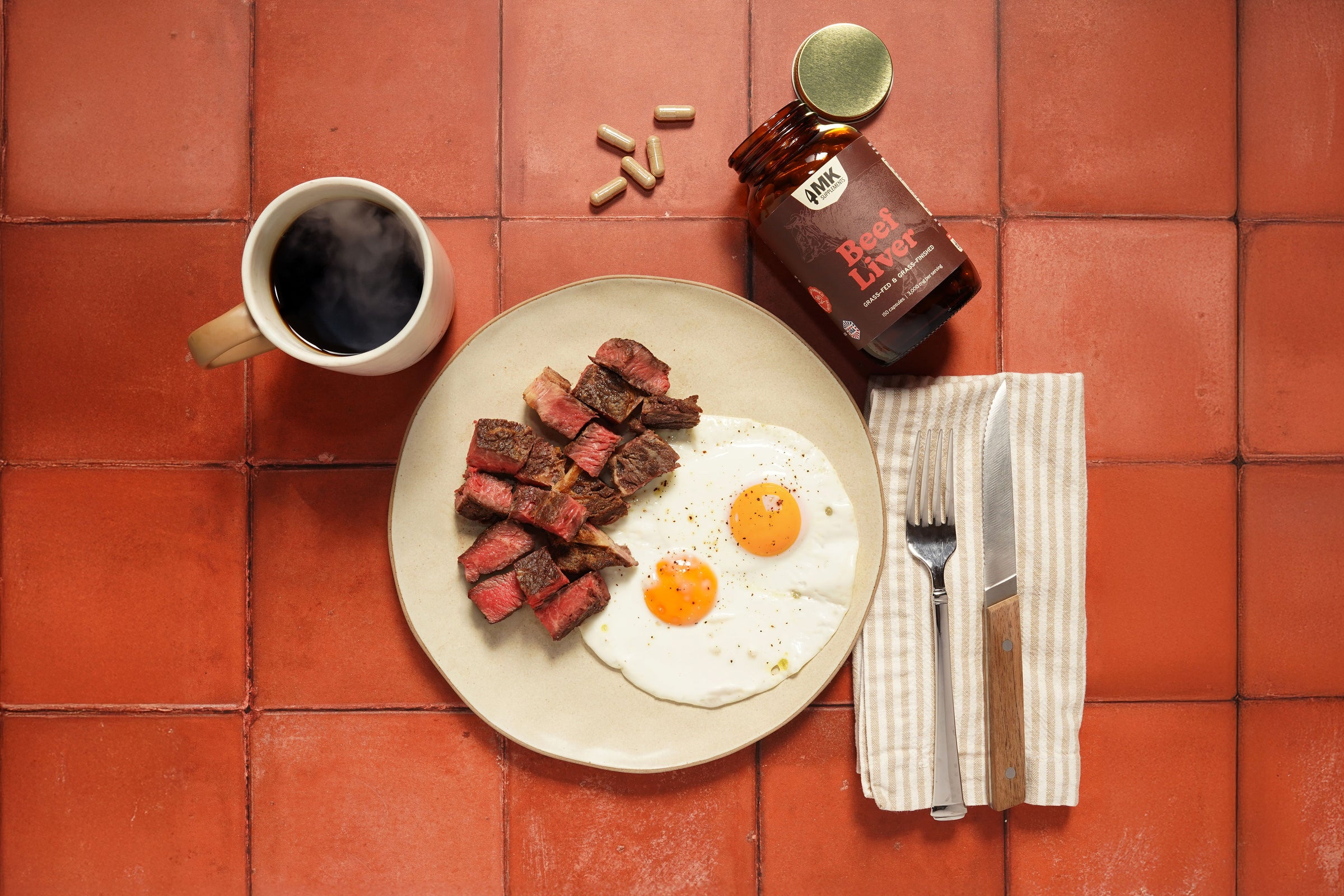 Breakfast setting with steak, eggs, coffee, and a bottle of beef liver supplement on a red tiled surface.